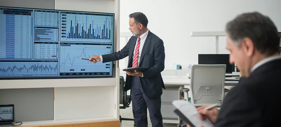 Business professional presenting data charts on a large screen in a modern office, while another colleague takes notes.