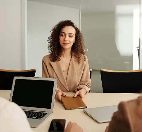 Young woman holding a notebook, seated at a meeting table with two laptops, engaging in a business discussion.