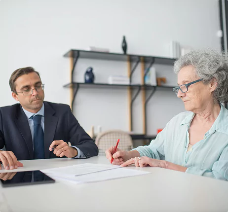 Elderly woman signing a document at a desk while a man in a suit attentively observes.