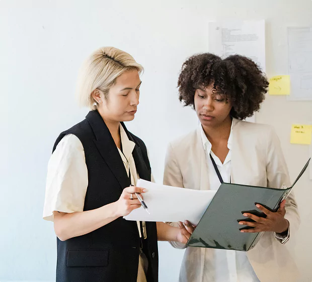 Two women in business attire review documents together in an office, standing near a wall with papers and sticky notes.