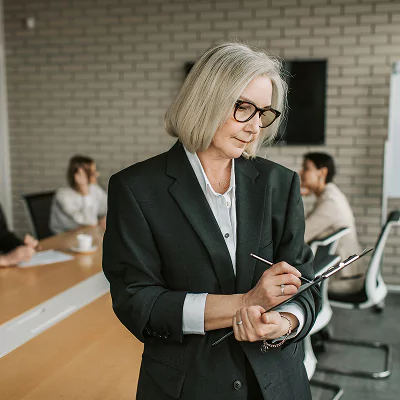 Senior woman in glasses and a black blazer writing on a clipboard during a business meeting, with a group of people discussing in the background.