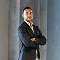 Confident businessman in a suit with arms crossed, standing in front of a grey wall