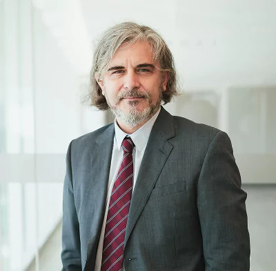Mature man with gray hair and beard wearing a dark suit and red-striped tie, standing in a modern office corridor.young woman with curly hair wearing a light blue blazer, standing with arms crossed in a bright, minimalistic office