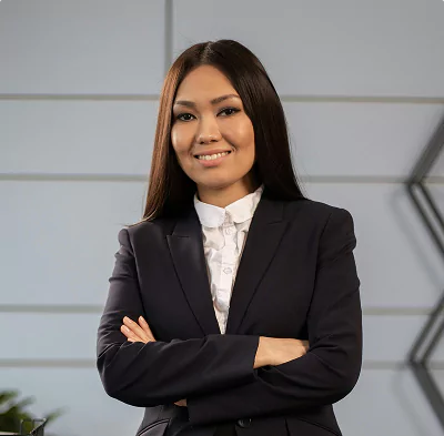 A woman in a dark blazer stands with arms crossed, smiling in a professional environment.