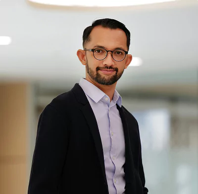 A man wearing glasses and a blazer smiles confidently in a modern office setting.