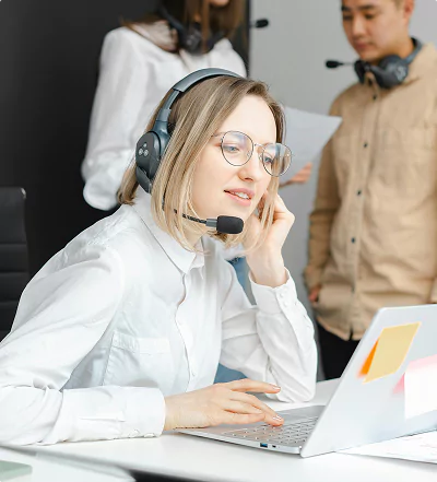 A woman with a headset works on a laptop in a support or call center setting.