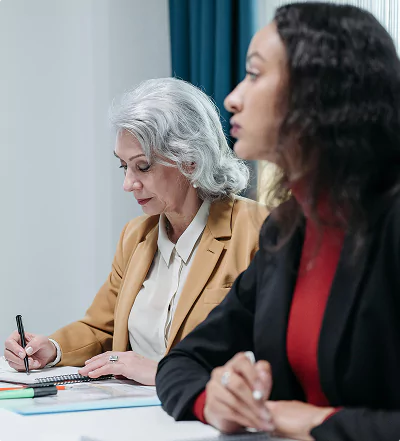 Two women in formal attire sit side by side in a meeting, one taking notes.
